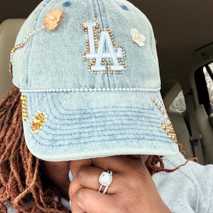 May include: Light blue denim baseball cap with the letters "LA" in white, embellished with gold and pearl beads, and floral accents. The cap is a stylish accessory.