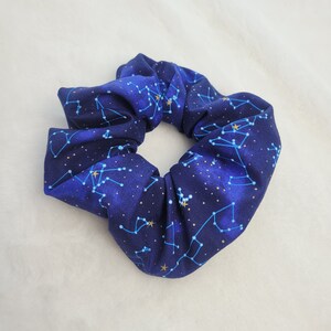 May include: A blue scrunchie with a constellation pattern. The scrunchie is made of a soft, stretchy fabric and features gold stars and constellations.