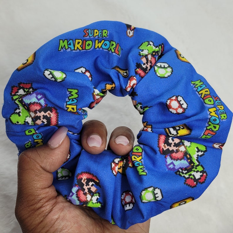Pu&ograve; includere: Scrunchie in tessuto blu con un motivo ripetuto di personaggi di Super Mario World e il testo "Super Mario World".