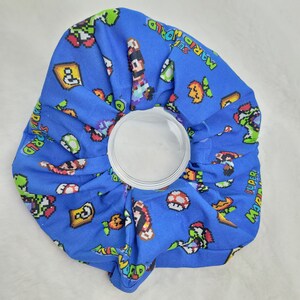 Pu&ograve; includere: Un impacco di ghiaccio in tessuto blu a tema Super Mario World. L'impacco di ghiaccio presenta vari personaggi e oggetti del videogioco Super Mario World, tra cui Mario, Luigi, Yoshi e i Goomba.