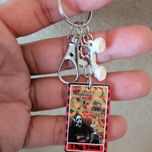 May include: A keychain with a rectangular charm featuring a horror movie theme. The charm has a red border and the words "HELLO SIDNEY" and "The Fool". A white telephone charm and a silver clasp and keyring are also attached.