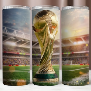 May include: A stainless steel tumbler with a gold-colored soccer trophy design. The trophy is a replica of the FIFA World Cup trophy. The background is a blurred image of a soccer stadium.
