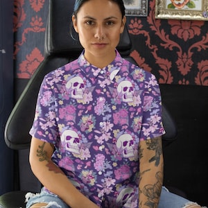 May include: A person wearing a purple short-sleeved button-down shirt with a floral and skull print. The shirt has a collar and a button closure. The person is sitting on a black leather chair.