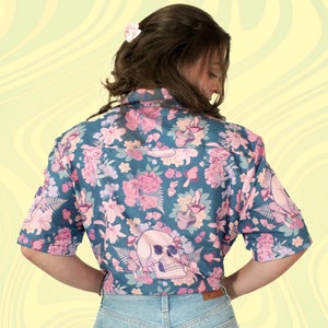 Puede incluir: Una camisa de botones de manga corta azul verdosa con estampado floral con flores rosas, setas y una calavera.