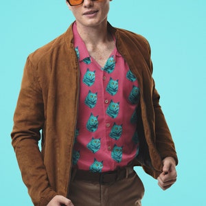 Puede incluir: Un hombre con una chaqueta de gamuza marrón sobre una camisa de botones rosa con estampado de gato azul. Lleva pantalones marrones y gafas de sol naranjas.