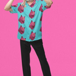 May include: A man wearing a light blue short-sleeved shirt with a pink cat print. He is wearing black pants and black shoes. The background is pink.
