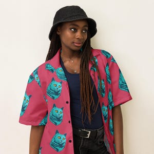 May include: A pink button-up shirt with a short sleeve and a blue cat print. The shirt has a collar and a button closure. The model is wearing a black bucket hat and black jeans.