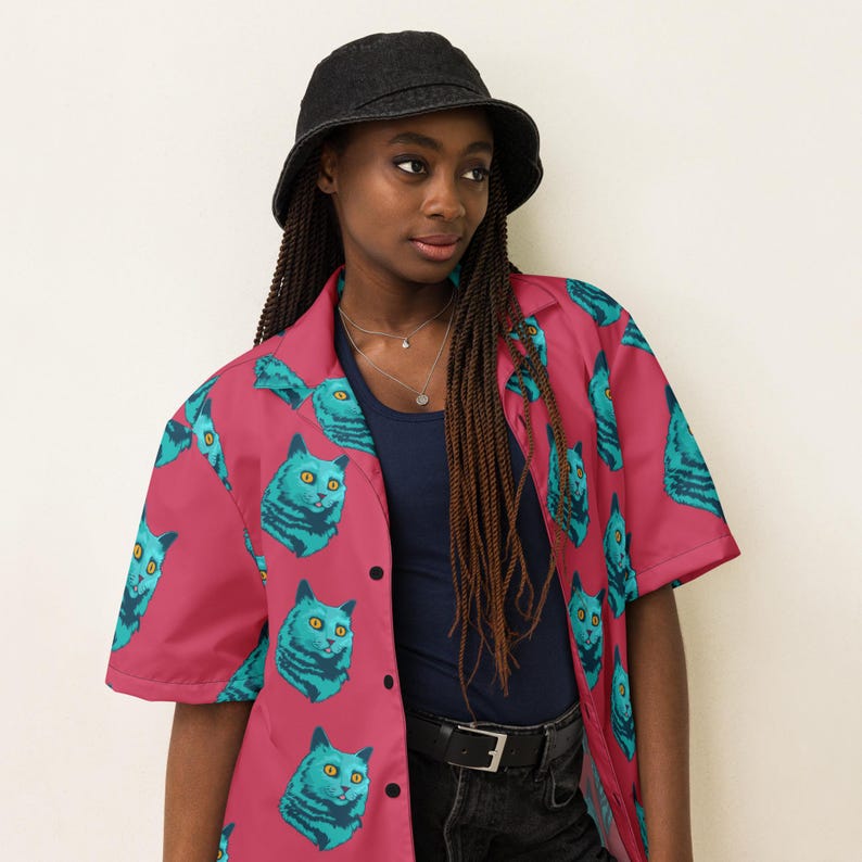 May include: A pink button-up shirt with a short sleeve and a blue cat print. The shirt has a collar and a button closure. The model is wearing a black bucket hat and black jeans.