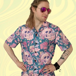 Puede incluir: Una camisa de botones de manga corta azul verdosa con estampado floral y de calaveras. La camisa tiene flores rosas, hojas verdes y calaveras blancas.