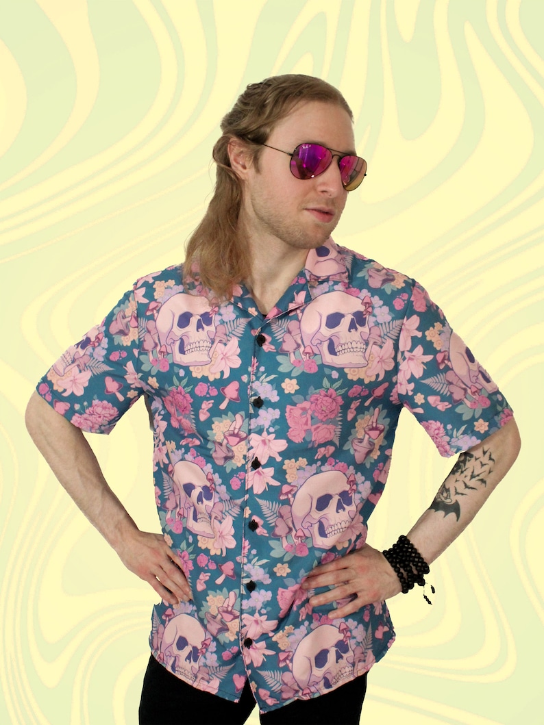 Puede incluir: Una camisa de botones de manga corta azul verdosa con estampado floral y de calaveras. La camisa tiene flores rosas, hojas verdes y calaveras blancas.