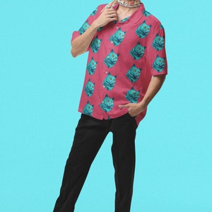 May include: A man wearing a pink short-sleeved shirt with a blue cat print and black pants. He is wearing glasses and a patterned scarf around his neck.