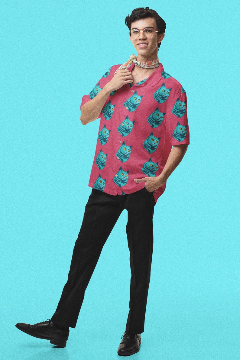 May include: A man wearing a pink short-sleeved shirt with a blue cat print and black pants. He is wearing glasses and a patterned scarf around his neck.