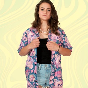 Puede incluir: Una camisa de botones con estampado floral azul y rosa con un dise&ntilde;o de calavera. La camisa est&aacute; abierta y deja ver un top negro sin mangas y unos pantalones cortos de mezclilla azul claro debajo.