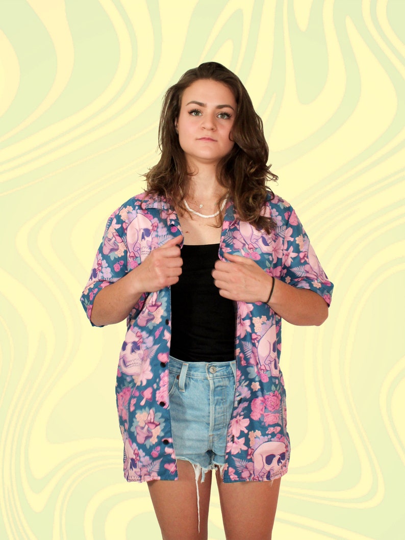 Puede incluir: Una camisa de botones con estampado floral azul y rosa con un dise&ntilde;o de calavera. La camisa est&aacute; abierta y deja ver un top negro sin mangas y unos pantalones cortos de mezclilla azul claro debajo.