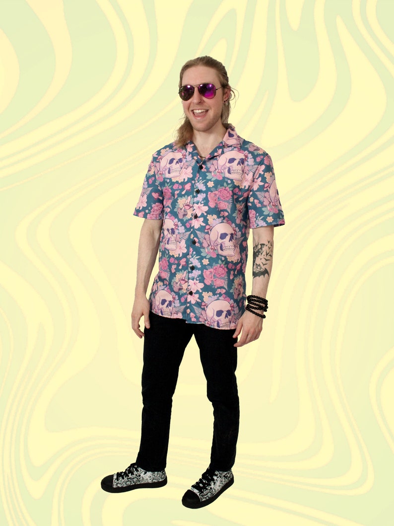 Puede incluir: Un hombre con una camisa de manga corta azul y rosa con botones, con un estampado de calavera y flores. Lleva pantalones negros y zapatillas negras y blancas. Tambi&eacute;n lleva gafas de sol.