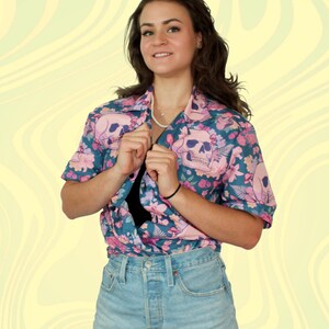 Puede incluir: Una mujer con una camisa de manga corta azul y rosa con estampado floral y calaveras y pantalones cortos de mezclilla.