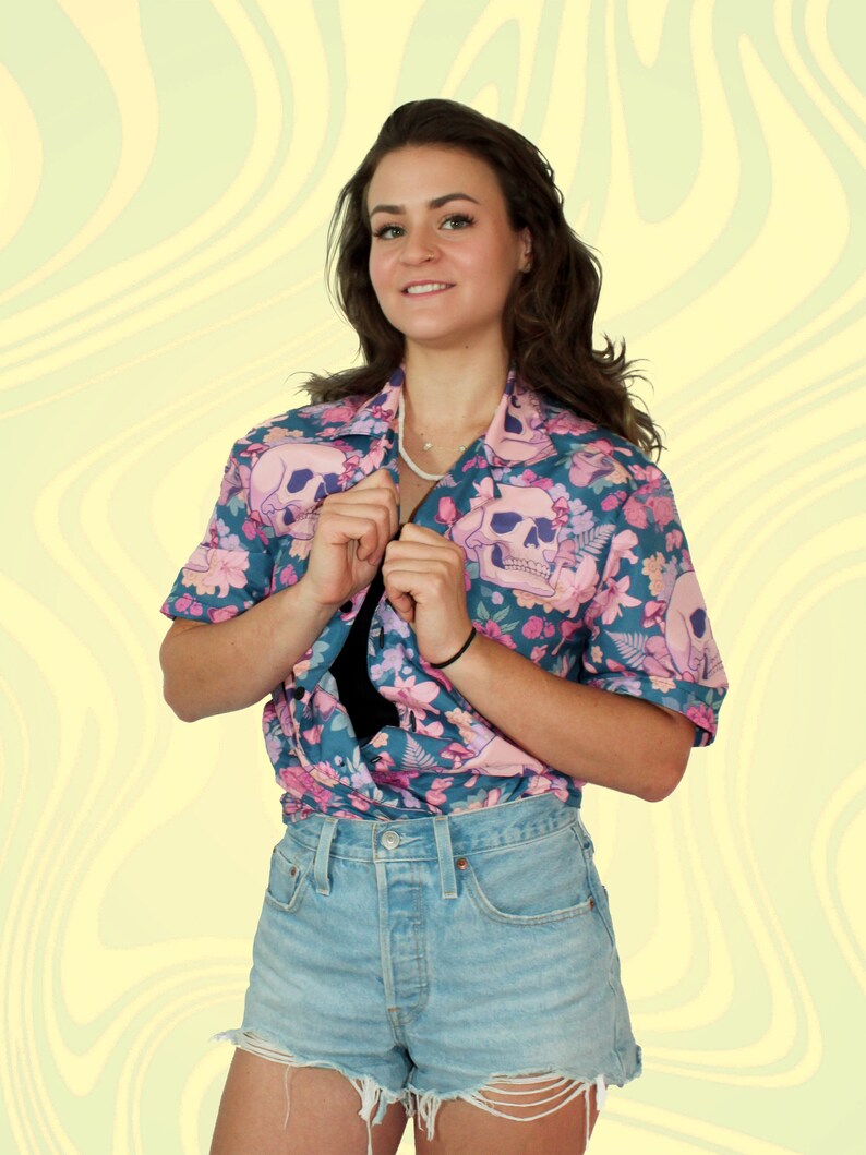 Puede incluir: Una mujer con una camisa de manga corta azul y rosa con estampado floral y calaveras y pantalones cortos de mezclilla.