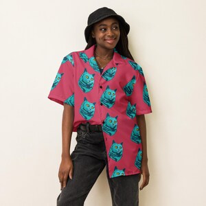 May include: A pink button-up shirt with a short sleeve and a cat print. The cat has blue fur and yellow eyes.