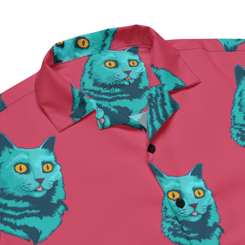 May include: A pink button-up shirt with a repeating pattern of blue cats with green eyes and a tongue sticking out. The cats are in a variety of poses, some are looking at the camera, some are looking away, and some are looking down. The shirt has a collar and a button-up front.