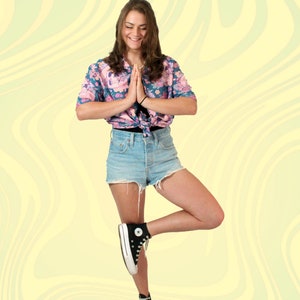 Puede incluir: Una mujer con una camisa de botones de estampado floral, pantalones cortos de mezclilla y zapatillas negras de ca&ntilde;a alta. Est&aacute; de pie sobre una pierna con las manos juntas frente a ella.