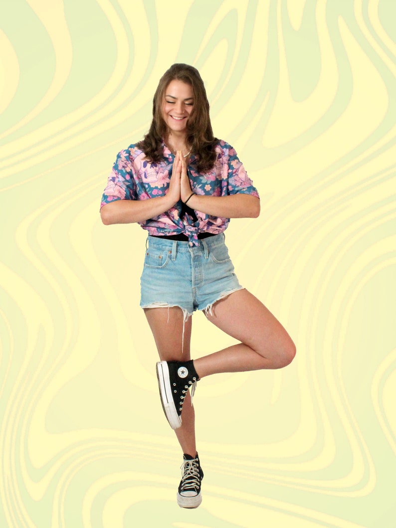 Puede incluir: Una mujer con una camisa de botones de estampado floral, pantalones cortos de mezclilla y zapatillas negras de ca&ntilde;a alta. Est&aacute; de pie sobre una pierna con las manos juntas frente a ella.