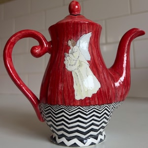 May include: A red ceramic teapot with a white angel illustration. The teapot has a black and white zigzag pattern at the base and a curved handle. The lid has a small red knob.
