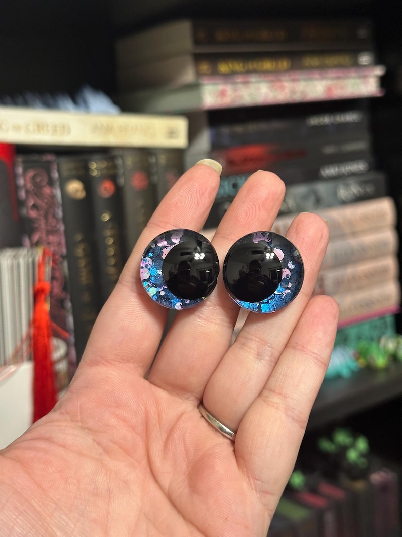 May include: A pair of black and blue glass eyes with iridescent glitter. The eyes have a black pupil and a white iris with pink and blue glitter.