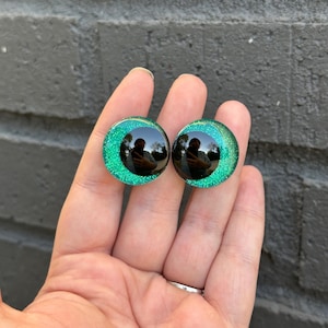 May include: Two green glitter round plastic eyes with black pupils. The eyes are about 1 inch in diameter.