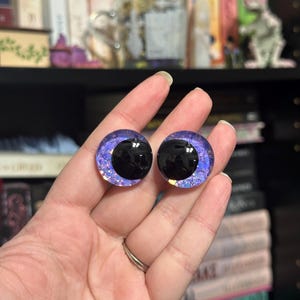 May include: Two purple and black glass eyes with glitter accents. The eyes are round and have a black pupil.