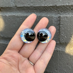 May include: Two iridescent blue and black glass eyes with a black pupil. The eyes are round and have a glittery finish.