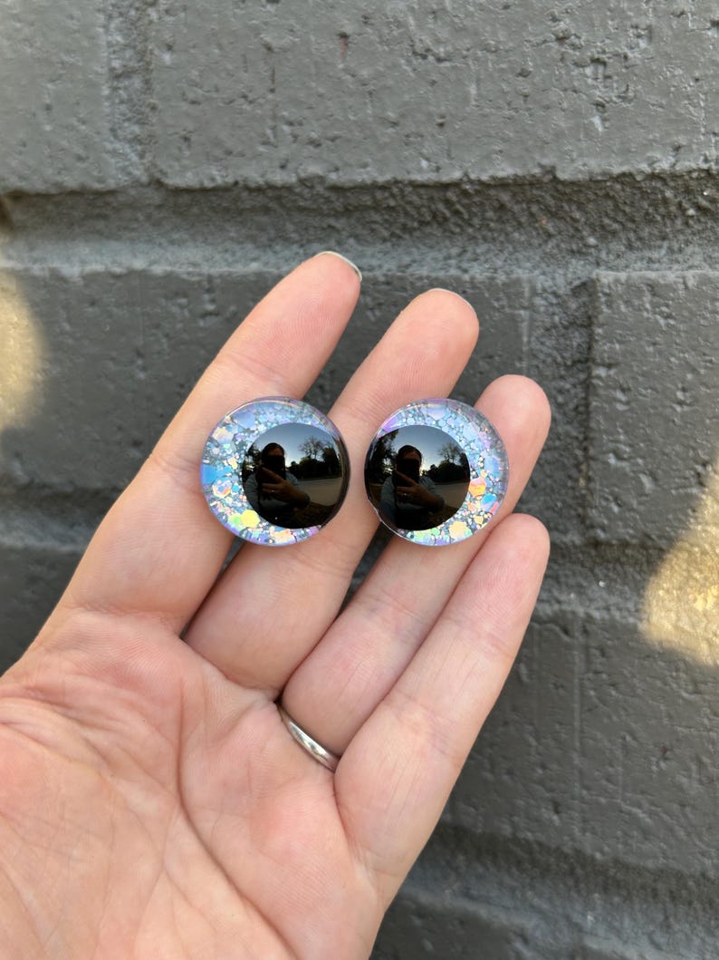 May include: Two iridescent blue and black glass eyes with a black pupil. The eyes are round and have a glittery finish.