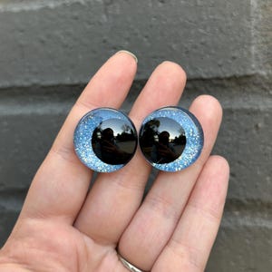 May include: Two blue glitter safety eyes with black pupils. The eyes are round and have a shiny finish.