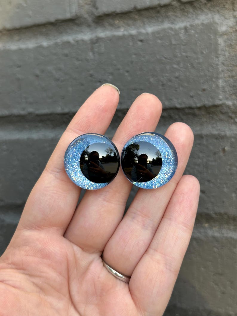 May include: Two blue glitter safety eyes with black pupils. The eyes are round and have a shiny finish.