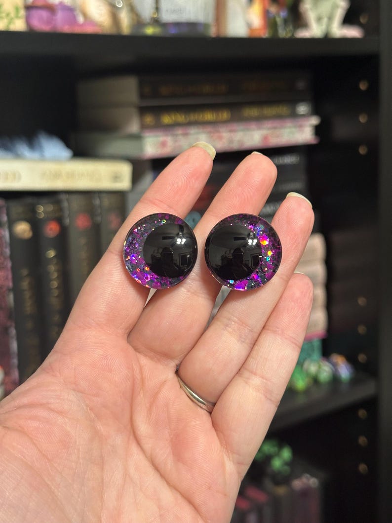 May include: Pair of black and purple glitter doll eyes with a black pupil. The eyes are round and have a glossy finish.