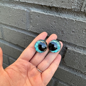 May include: Two round, black, plastic eyes with a turquoise glitter finish. The eyes are about 1 inch in diameter.
