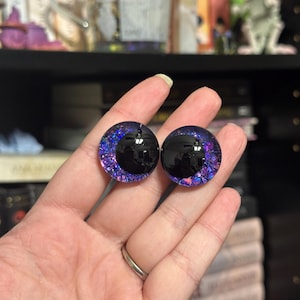 May include: Two round, black-pupil, glass eyes with a purple and blue glitter rim. The eyes are approximately 2.5 cm in diameter.