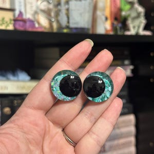 May include: A pair of black and green glitter doll eyes with a black pupil. The eyes are round and have a shiny finish.