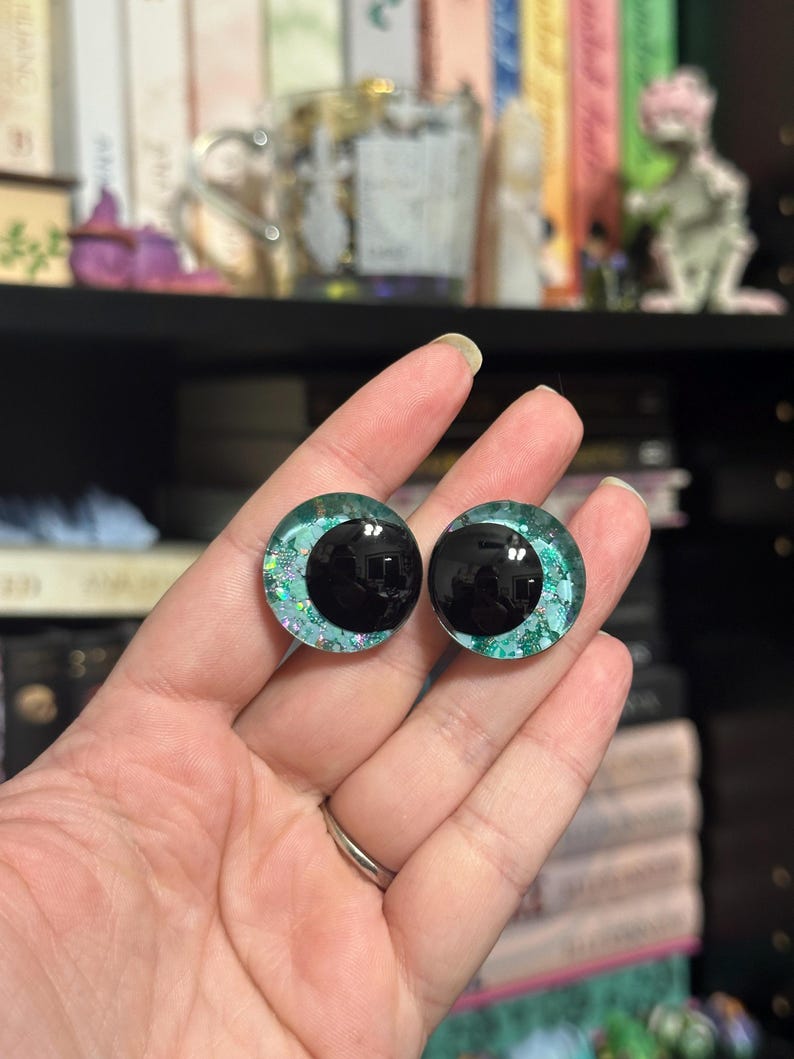 May include: A pair of black and green glitter doll eyes with a black pupil. The eyes are round and have a shiny finish.