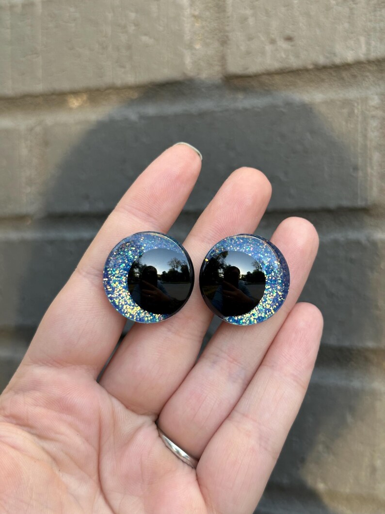 May include: Two black and blue glitter safety eyes with a black pupil. The eyes are round and have a shiny finish.