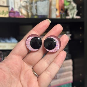 May include: Two pink and black plastic doll eyes with glitter accents. The eyes are round and have a black pupil.