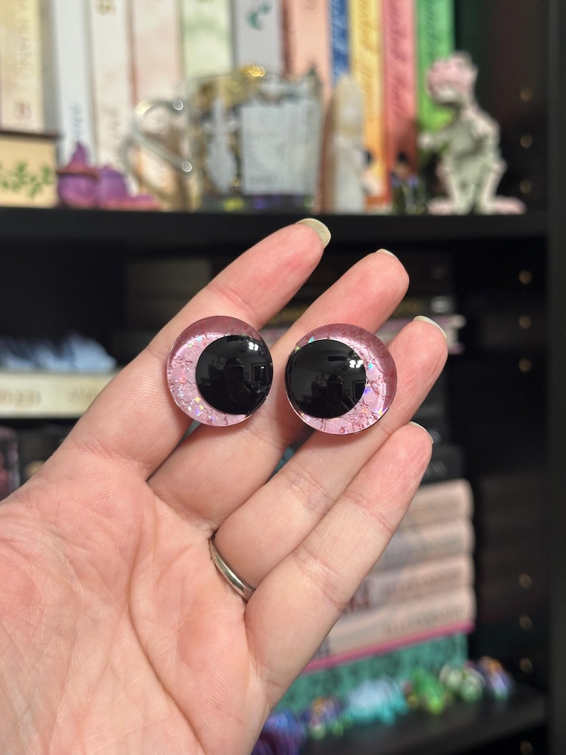 May include: Two pink and black plastic doll eyes with glitter accents. The eyes are round and have a black pupil.