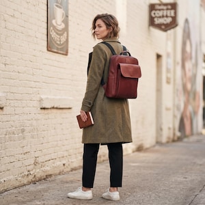 May include: A person wearing an olive green coat, black trousers, and white trainers, carrying a brown leather backpack and a small brown notebook. The backpack has a flap closure. A coffee shop sign is visible.