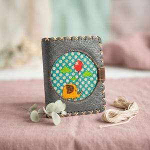 May include: A gray leather wallet with a stitched border and a brown leather clasp. The front features a circular design with a cartoon elephant holding a red balloon against a blue polka dot background.