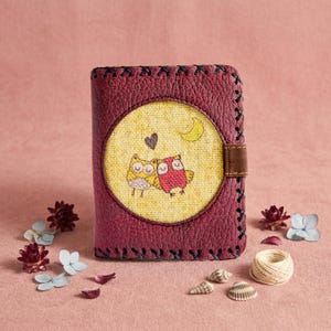 May include: A burgundy leather wallet with a stitched border and a brown leather strap. The front features a circular design with two cartoon owls, a heart, and a crescent moon. Decorative flowers, shells, and twine are scattered around the wallet.