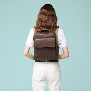 May include: A brown leather backpack with a rectangular design and a front pocket. The backpack has a top handle and adjustable straps. The person is wearing a white t-shirt and white trousers. The background is a light blue.