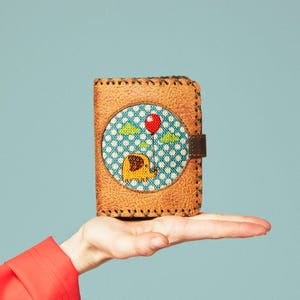 Puede incluir: Una cartera de cuero marrón con un borde cosido. La parte delantera presenta un diseño bordado circular de un elefante amarillo sosteniendo un globo rojo sobre un fondo a cuadros azul y blanco. Una correa de cuero marrón asegura la cartera.