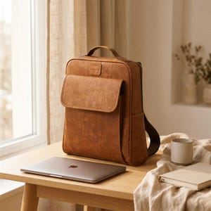 May include: A brown leather backpack with a front flap and top handle sits on a wooden table. A closed laptop and a small ceramic mug are also on the table. The backpack has a textured appearance and a rectangular shape.