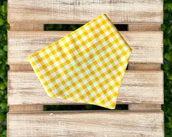 May include: A yellow and white gingham patterned pet bandana. The bandana is folded and placed on a rustic wooden crate. The background is a lush green plant.