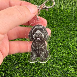 May include: A clear acrylic keychain featuring a black and white illustration of a seated dog with a happy expression. The keychain has a silver-colored clasp for easy attachment to keys or bags. The dog's fur is detailed, and the overall design is playful.