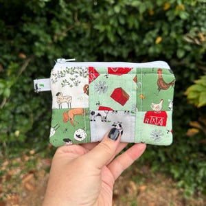May include: A small, patchwork fabric zippered pouch featuring farm animal and barnyard-themed patterns in green, white, and red. The pouch is held in a hand against a blurred green background. The design includes cows, chickens, and barns.